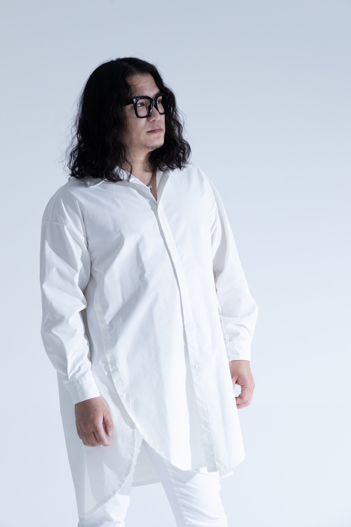 白いシャツを着て立つ華僑J。メガネをかけ、横を向いている。